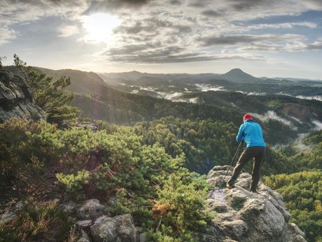 Photographer in blue sweatshirt working with mirror camera and tripod on peak of rock. Dreamy foggy landscape.