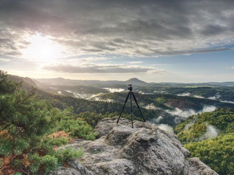 Tripod on peak ready for photography. Sharp autumn rocky peaks with hills and mist in deep valley 