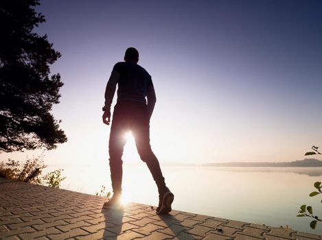 Tall man with sunglasses, red baseball cap and blue black sportswear is running and exercising on sea bridge. Sport active man.