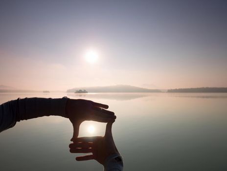 Man hand makes shadow symbols  to Sun. Misty daybreak at a beautiful peaceful lake.