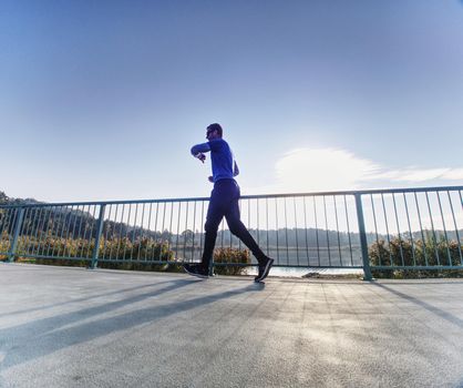 Tall man with sunglasses, red baseball cap and blue black sportswear is running and exercising on sea bridge. Sport active man.