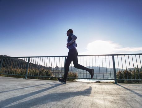 Man is running quickly on the shore bridge. Silhouette of active man exercising and stretching on the lake street. Healthy lifestyle. Alone young fitness man.