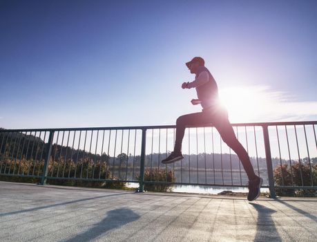 Side shot of healthy middle age man running on lake promenade in sunny morning. Male runner sprinting outdoors. Down angle view