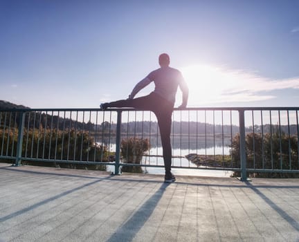 Man stretching legs before going  run outside within sunny morning. Warming up muscles. Outdoor exercising on lake bridge.