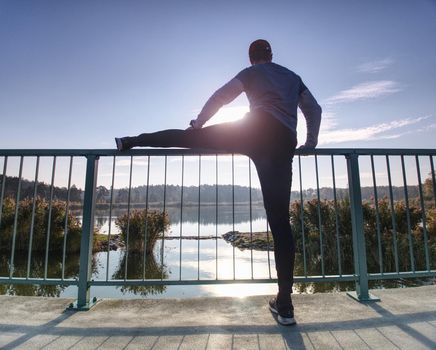 Man is stretching legs in break from run.  The Sun is outlining man body. Outdoor exercising on smooth concrete ground on lake bridge.