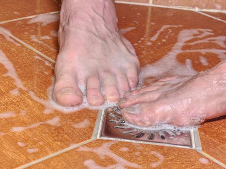 Couple of naked feet in shower. Detailed foot toes, floor tilles, stainless sink. Feet washing in bathroom with shampoo foam.