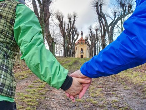 Couple tourists hold hands and walking through alley in park, old chapel at the end