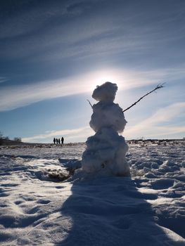 Real icy snowman standing in winter landscape. Hot spring sun and some people walk at horizon