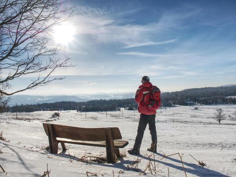 Man tourist is resting at wooden bench in winter landscape. Hiker with red  backpack