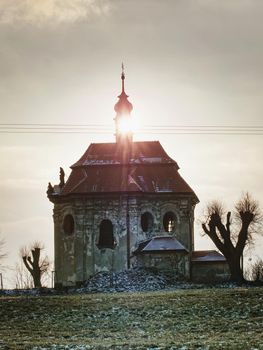 Forgotten chapel between old willows and lime trees on hill. Cold sun behind of bell tower