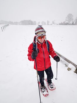 Sports body woman walk quickly with red hobby snowshoes. girl walk in black leggings and warm red jacket, knitted hat and orange sunglasses. 