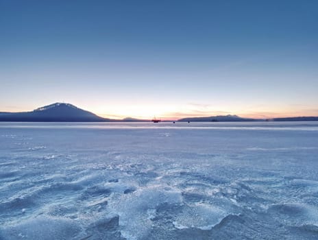 Breathtaking sunset with golden and pink clouds over frozen lake. Footprints across snow covered lake. Winter season greeting card background with copy space.