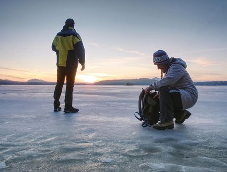 Traveler stay on ice of frozen sea. Woman with backpack and long warm jacket. Footprints in ice of laggon. Orange sun is rissing above hilly horizon.