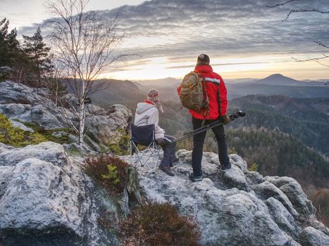 Partners enjoy photographying in wild nature. Nature photographers with big camera on tripod stay on summit rock. 