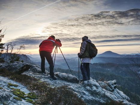 Couple hiking photographers on view point above forest, taking photos and looking on sunset