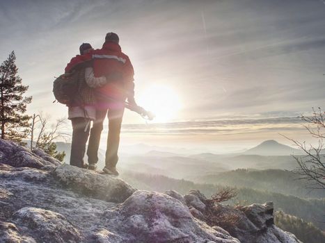 Couple of hikers look down into fogy valley. Photographer stay on cliff and takes photos. Dreamy fogy landscape blue misty sunrise in a beautiful valley below