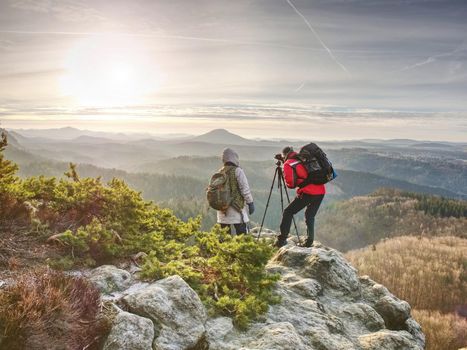 Nature photographer with tripod and camera on cliff and thinking. Dreamy fogy landscape orange misty sunrise in a beautiful mountains