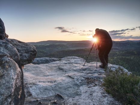 Professional photographer stay with tripod on cliff and thinking.  Foggy landscape spring orange pink misty sunrise in beautiful valley below.