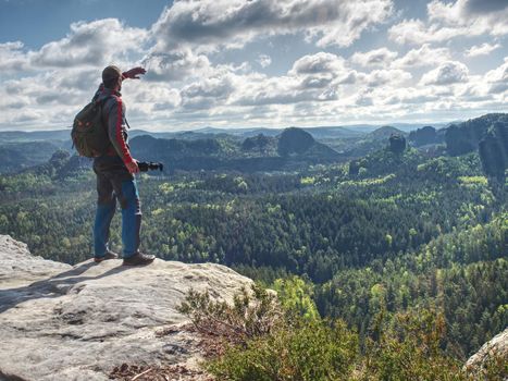 Photographer with heavy backpack and tripod in hand on rocky cliff and watching down to deep valley