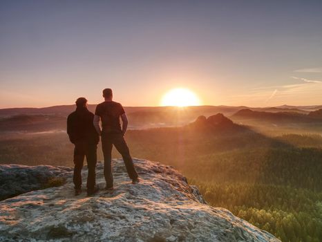 Hikers and friends stay on cliff within dreamy daybreak and thinking. Dreamy fogy landscape blue misty sunrise in a beautiful valley below