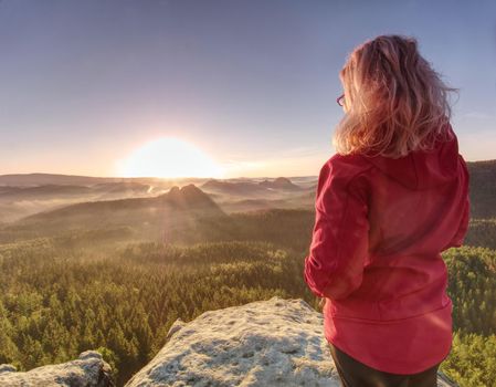 Girl watch Sun rising at horizon. Colorful spring valley with stripped mist and stiicking out treetops.