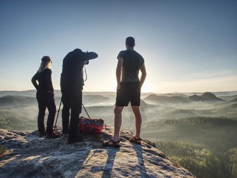 Three friends photographers discuss and taking photo against sunset sky from sticking out rock above misty pandscape. Photographers backpacker and hikers style. Concept of active leisure.