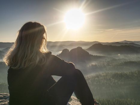 Blond woman sitting on the edge of the mountain cliff against beautiful mountains peak. Travel and lifestyle concept