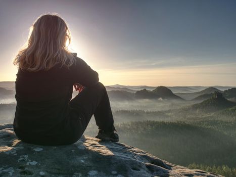 Blond woman sitting on the edge of the mountain cliff against beautiful mountains peak. Travel and lifestyle concept