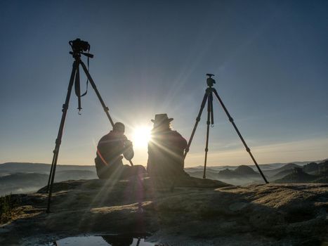 Creative artists stay at own cameras on tripods.  Hikers and photo enthusiasts work together on cliff and thinking