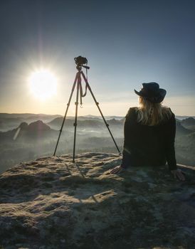 Blond hair woman hiker sit and thinking about taking photo with digital camera at mountain peak. Amazing view from cracked cliff edge of sandstone rock.