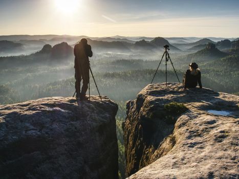 Silhouette of three art photographers overlooking misty valley to sun.  Female and male artists in trendy outdoor clothes posing at professional cameras with a tripods at edge