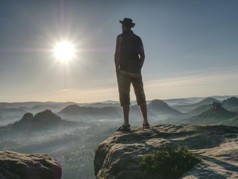 Tall man in shorts wearing a cowboy hat shielding his face from the sun, watching over large valley bellow sharp rocky shields.