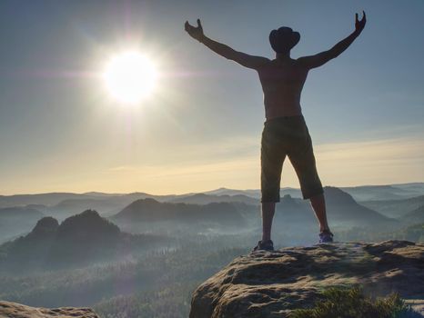 Silhouette of tall naked hiker wearing cowboy hat. Guy looking over large valle from sharp mountain shield.