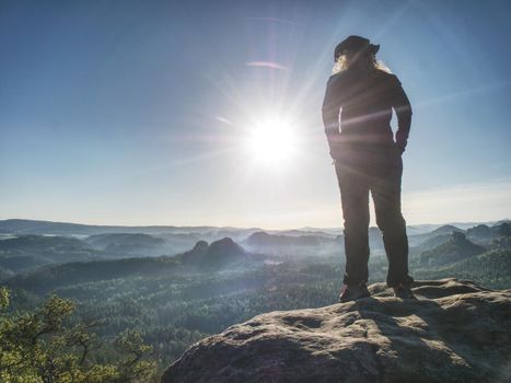 Hiker girl with leather cap stay with hands in pocket on cliff edge above misty valley. Marvelous autumn morning in pure nature