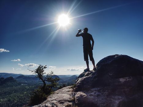 Traveler on rock drinking bottled water from transparent plastic bottle. Morning sunlight with sun flares