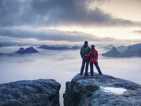 Lovely heterosexual Couple looking at far sunrise in heavy clouds. The dark night in foggy mountains ends.