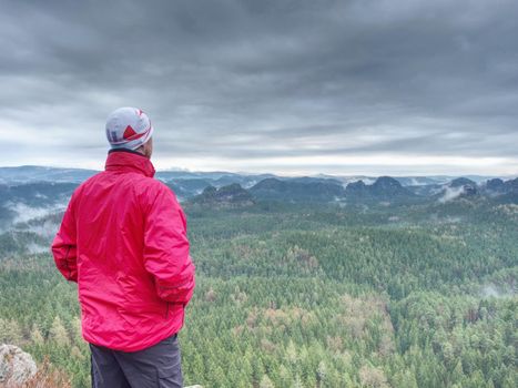 Sportsman in red with hands in pockets stand on the peak in rock empires park and watching over the misty and foggy morning valley to Sun.