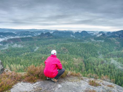 Sportsman in red with hands in pockets stand on the peak in rock empires park and watching over the misty and foggy morning valley to Sun.