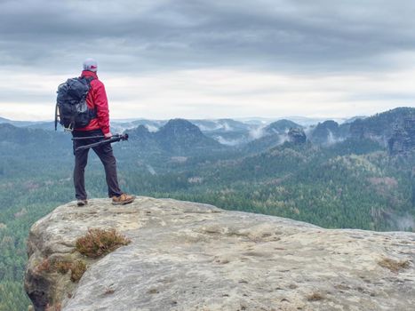 Alone hiker in warm red  jacket stand on peak of sandstone rock  and waiting for Sun. Misty and foggy morning valley