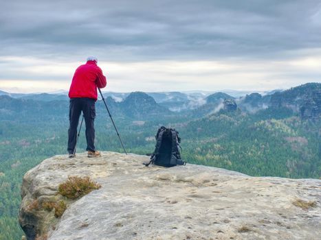 Outdoor photographer with tripod and camera on rock thinking. Mist in valley bellow. 