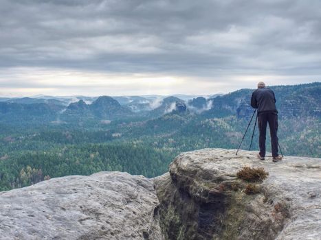 Photographer check display of camera on tripod. Man stay on cliff and takes photos talk friends. Autumn beautiful misty landscape misty sunrise at horizon. Men in mountains