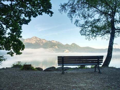 Bench under a tree on a lake shore. Mountains at background. Take a rest near blue green lake.  Beautiful mystical lake.