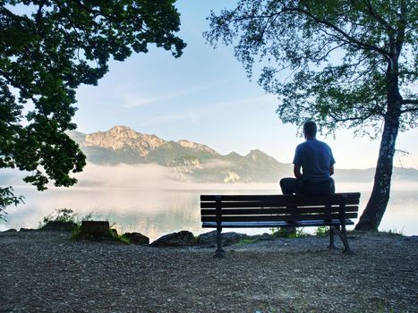 Man sit lone on a bench in park next lake. Mountain range with amazing view over smooth lake surface.