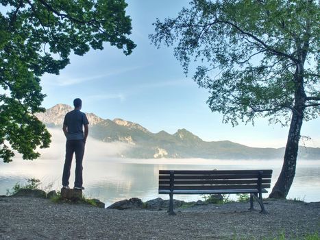 Tall man walks at bench on lake shore and looks at the sports boat on the level.