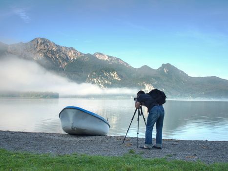 Man hiker is taking photo of ship at mountain lake shore. Silhouette at fishing paddle boat at lake coast.