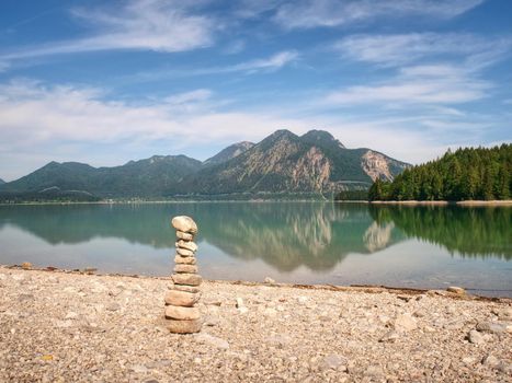 Pyramid of flat stones on a pebbly lake beach, the mountains mirroring in smooth water level.