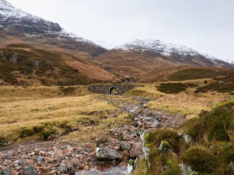 Cold foggy day in spring Higland mountains in Scotland.  Snowy cone of mountain in heavy clouds. Dry grass and heather bushes on stream banks. 