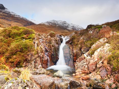 Rapids in small waterfall on stream, Higland in Scotland an early spring day. Snowy cone of mountain in clouds. Dry grass and heather bushes on banks