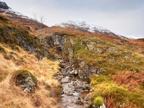 Rapids in small waterfall on stream, Higland in Scotland an early spring day. Snowy cone of mountain in clouds. Dry grass and heather bushes on banks