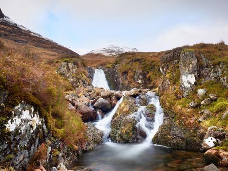 Rapids in small waterfall on stream, Higland in Scotland an early spring day. Snowy cone of mountain in clouds. Dry grass and heather bushes on banks